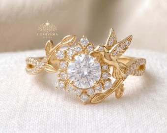 May include: A gold-colored ring featuring a central, round, clear gemstone surrounded by smaller, clear gemstones. The ring has a floral and bird design with leaves and a bird on either side of the central stone. The ring band has a twisted design.