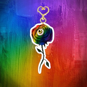 Peut inclure: Un porte-clés en forme de rose aux couleurs de l'arc-en-ciel avec un globe oculaire humain au centre des pétales. La rose possède une tige verte avec des épines et des feuilles, fixée à un fermoir en métal doré en forme de cœur.