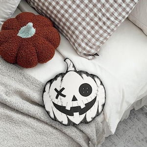 May include: A white pumpkin-shaped throw pillow featuring a black hand-drawn style illustration of a jack-o'-lantern face with a button eye, an X-shaped eye, and stitched mouth details, positioned next to a textured rust-colored pumpkin pillow.