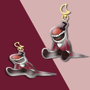 Puede incluir: Dos colgantes de acrílico que muestran un personaje con sombrero de copa rojo, abrigo rojo y una boca ancha llena de dientes blancos afilados. El personaje tiene un ojo oscuro cosido y lleva un chaleco blanco. Cada colgante incluye un cierre dorado en forma de luna.