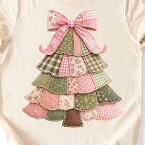 May include: A cream-coloured short-sleeve t-shirt featuring a central graphic of a Christmas tree constructed from various patches of fabric in shades of pink, green, and white, topped with a large pink and white plaid fabric bow.