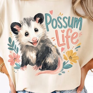 May include: A cream-colored t-shirt featuring a graphic illustration of a smiling opossum surrounded by colorful flowers and leaves. The text "Possum Life" is printed in teal and coral script above the animal, accented by small hearts.
