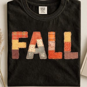 Peut inclure: Un t-shirt noir à manches courtes avec le mot FALL écrit en grandes lettres style bloc sur la poitrine. Chaque lettre est composée de divers motifs de tissu en patchwork dans des tons orange, marron et crème, avec des coutures visibles.