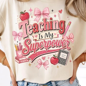 May include: A cream-colored t-shirt featuring a graphic print with the text Teaching Is My Superpower. The design includes a red apple, pink bows, a pencil holder with pencils, a stack of books labeled TEACH, and a notebook labeled Lesson Plans.