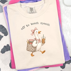 Op de afbeelding: Een crèmekleurig t-shirt met een afbeelding van een witte gans met een roze gestrikte strik en een halsdoek. De gans draagt een tas met luipaardprint met potloden en een notitieboekje en houdt een ijskoffie vast. De tekst luidt off to teach speech.