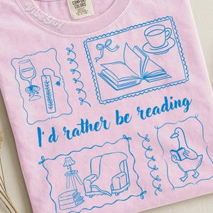 Peut inclure: T-shirt rose clair avec un motif bleu. Le motif comprend des illustrations d'un verre à vin, d'un livre, d'une tasse à thé, d'une oie lisant et d'un fauteuil. Le texte "I'd rather be reading" est également présent.