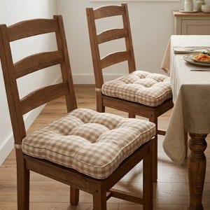 Puede incluir: Dos sillas de comedor de madera con cojines de asiento a cuadros marrones claros y blancos. Las sillas tienen un diseño de respaldo de escalera y están cerca de una mesa preparada para una comida. La habitación tiene una paleta de colores neutros.
