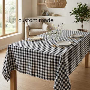 Könnte beinhalten: Eine rechteckige Tischdecke mit schwarz-weißem Vichy-Karo-Muster auf einem Holztisch. Der Tisch ist mit weißen Keramiktellern, goldfarbenem Besteck und klaren Glasvasen mit kleinen weißen und violetten Blumen gedeckt.