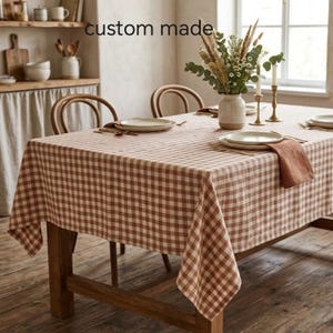May include: A rectangular dining table covered with a brown and white chequered tablecloth. The table is set with white plates, a folded brown napkin, and a central ceramic vase containing dried flowers. The text custom made is visible.