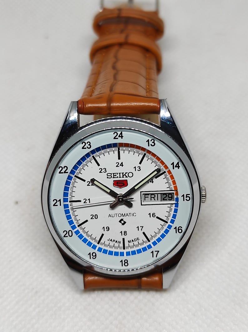 May include: A silver-cased Seiko automatic watch with a white face, featuring black hands and markers. The watch has a brown leather strap and displays the day and date. The face includes the text "SEIKO" and "AUTOMATIC".