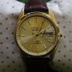 K&ouml;nnte beinhalten: Eine goldfarbene Citizen Automatik-Armbanduhr mit braunem Lederarmband. Das Zifferblatt zeigt Tag und Datum, mit "FRI 8" sichtbar. Die Uhr hat eine goldene L&uuml;nette und ein goldfarbenes Zifferblatt mit gr&uuml;nen und schwarzen Akzenten.