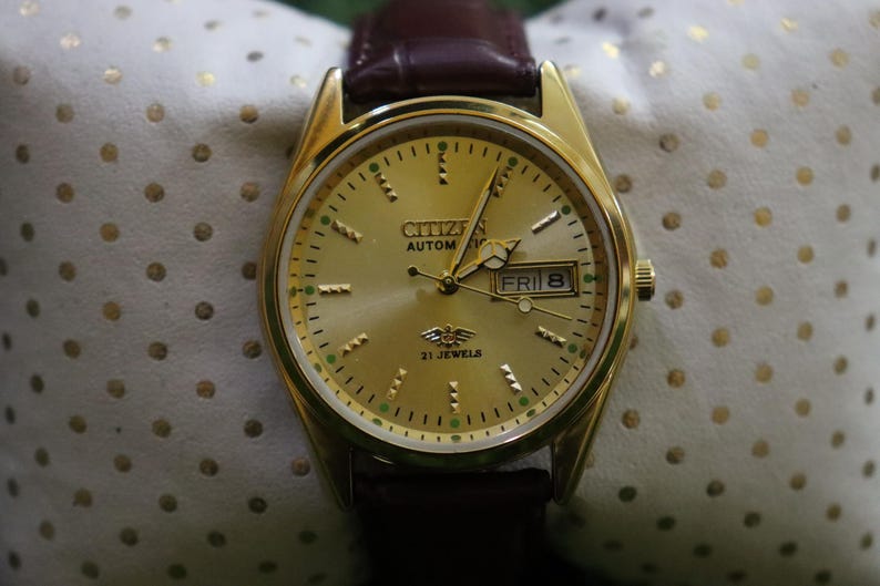 K&ouml;nnte beinhalten: Eine goldfarbene Citizen Automatik-Armbanduhr mit braunem Lederarmband. Das Zifferblatt zeigt Tag und Datum, mit "FRI 8" sichtbar. Die Uhr hat eine goldene L&uuml;nette und ein goldfarbenes Zifferblatt mit gr&uuml;nen und schwarzen Akzenten.