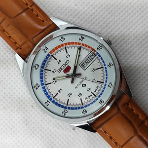 May include: A silver Seiko automatic watch with a white face, featuring black hands and markers, and a day-date display. The watch has a brown leather strap. The face has blue and orange accents, and the text "SEIKO AUTOMATIC" is visible.