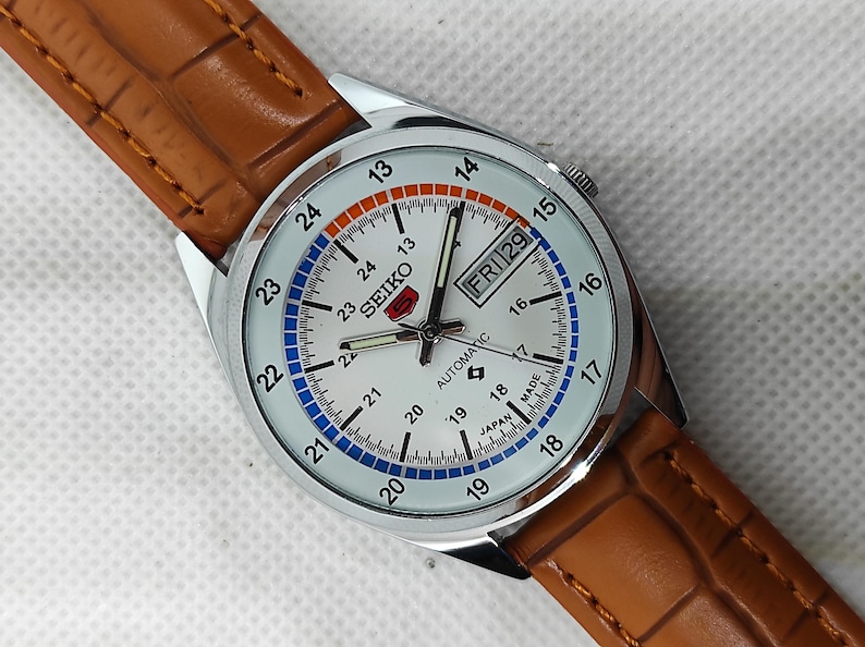May include: A silver Seiko automatic watch with a white face, featuring black hands and markers, and a day-date display. The watch has a brown leather strap. The face has blue and orange accents, and the text "SEIKO AUTOMATIC" is visible.