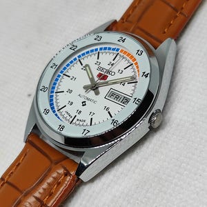 May include: A silver-toned Seiko automatic watch with a white face, day/date display, and a brown leather strap. The watch face features blue and orange accents, with the Seiko logo and the text "AUTOMATIC" visible. The watch is made in Japan.