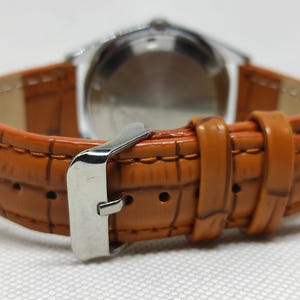 May include: Close-up of a watch with a brown leather strap. The strap has a textured, crocodile-like pattern and a silver buckle. The watch face is visible in the background.