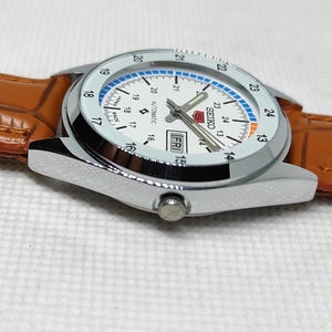 May include: A silver Seiko automatic watch with a white face, blue and orange accents, and a brown leather band. The watch displays the day and date, with the day reading "FRI". The watch has a stainless steel case.