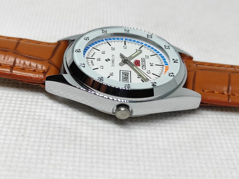 May include: A silver Seiko automatic watch with a white face, blue and orange accents, and a brown leather band. The watch displays the day and date, with the day reading "FRI". The watch has a stainless steel case.