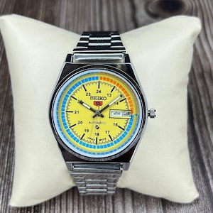 Könnte beinhalten: Eine Vintage Seiko 5 Automatik-Uhr mit gelbem Zifferblatt, blauen und orangefarbenen Akzenten und einem Edelstahlarmband. Das Zifferblatt zeigt Tag und Datum mit dem Text "SEIKO 5 AUTOMATIC".