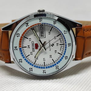 May include: A Seiko automatic watch with a white face, silver bezel, and brown leather band. The watch face displays the day and date, with a 24-hour clock. The band is a light brown color.
