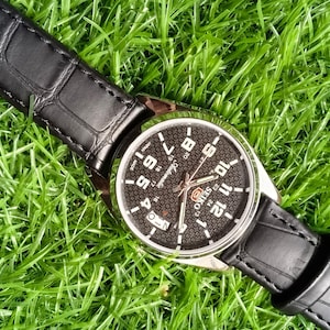 Puede incluir: Un reloj de pulsera negro con una esfera negra texturizada y una caja plateada. El reloj tiene números y agujas blancas, y una correa de cuero negro con un patrón de cocodrilo. El reloj está sobre hierba verde.