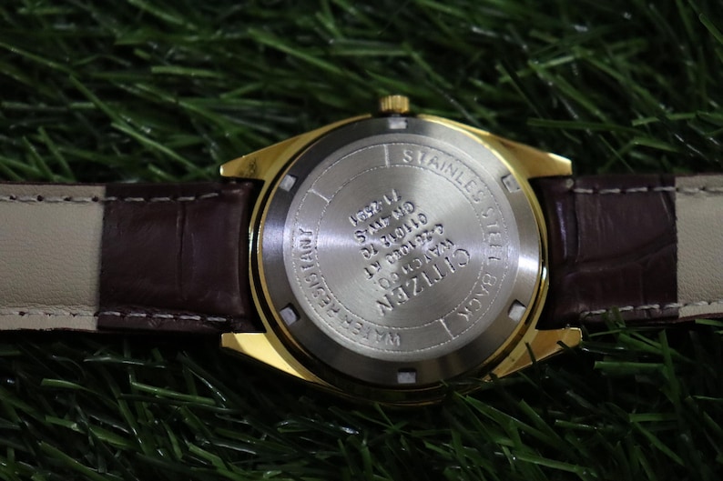 K&ouml;nnte beinhalten: Nahaufnahme einer goldfarbenen Citizen-Uhr mit braunem Lederarmband. Der Uhrenboden ist aus Edelstahl und tr&auml;gt die Aufschrift "WATER RESISTANT" und "CITIZEN WATCH CO". Die Uhr ist vor einem gr&uuml;nen Hintergrund.