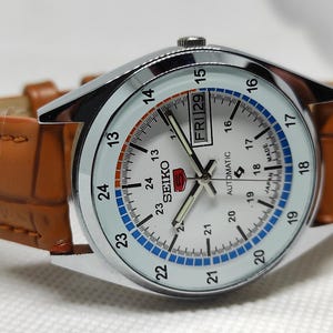 May include: A silver-toned Seiko 5 automatic watch with a white face, featuring a day and date display. The watch has a brown leather band. The watch face displays the time with black hands and numerals.