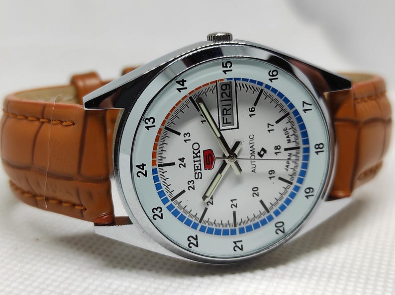 May include: A silver-toned Seiko 5 automatic watch with a white face, featuring a day and date display. The watch has a brown leather band. The watch face displays the time with black hands and numerals.