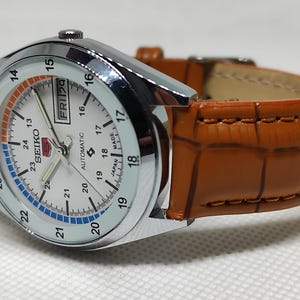 May include: A silver-toned Seiko automatic wristwatch with a white face, featuring black numerals and a day-date display. The watch has a brown leather band with a crocodile pattern. The watch face has orange and blue accents.