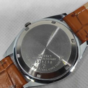 May include: Close-up of a Seiko watch back with a brown leather strap. The watch back is stainless steel with the text "SEIKO WATER RESISTANT 335718 TL STAINLESS STEEL 6309-5820".