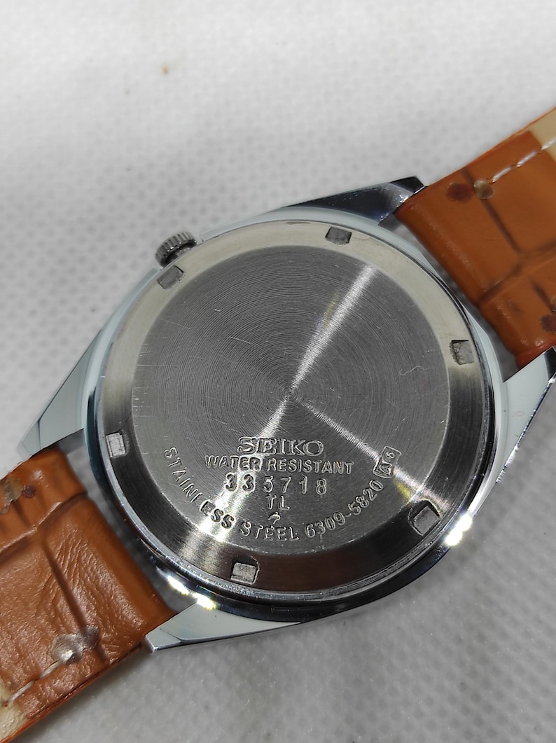 May include: Close-up of a Seiko watch back with a brown leather strap. The watch back is stainless steel with the text "SEIKO WATER RESISTANT 335718 TL STAINLESS STEEL 6309-5820".