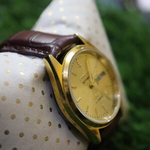 K&ouml;nnte beinhalten: Eine goldfarbene Armbanduhr mit braunem Lederarmband. Das Zifferblatt zeigt den Tag und das Datum an. Die Uhr liegt auf einem wei&szlig;en Tuch mit goldenen Punkten. Der Hintergrund ist ein verschwommenes Gr&uuml;n.