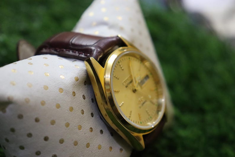 K&ouml;nnte beinhalten: Eine goldfarbene Armbanduhr mit braunem Lederarmband. Das Zifferblatt zeigt den Tag und das Datum an. Die Uhr liegt auf einem wei&szlig;en Tuch mit goldenen Punkten. Der Hintergrund ist ein verschwommenes Gr&uuml;n.