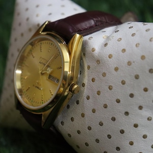 K&ouml;nnte beinhalten: Eine goldfarbene Armbanduhr mit braunem Lederarmband liegt auf einem wei&szlig;en Kissen mit goldenen Punkten. Das Zifferblatt zeigt den Tag und das Datum an. Die Uhr ist ein Automatikmodell.