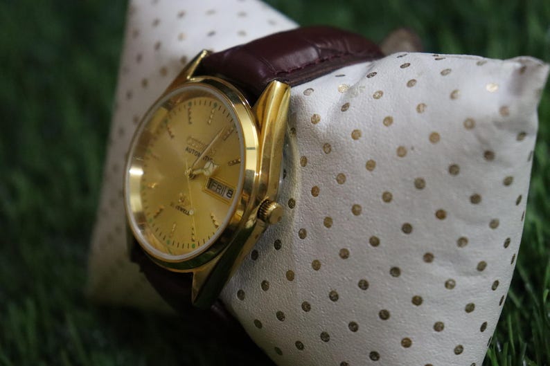K&ouml;nnte beinhalten: Eine goldfarbene Armbanduhr mit braunem Lederarmband liegt auf einem wei&szlig;en Kissen mit goldenen Punkten. Das Zifferblatt zeigt den Tag und das Datum an. Die Uhr ist ein Automatikmodell.