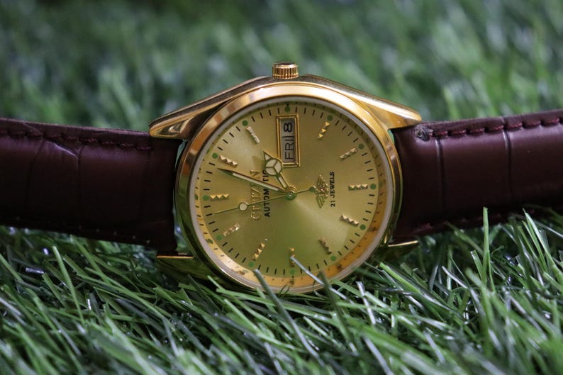 K&ouml;nnte beinhalten: Eine goldfarbene Citizen Automatik-Armbanduhr mit braunem Lederarmband. Das Zifferblatt der Uhr ist goldfarben mit schwarzen Stundenmarkierungen und Zeigern. Tag und Datum werden auf 3 Uhr angezeigt. Die Uhr liegt auf gr&uuml;nem Gras.