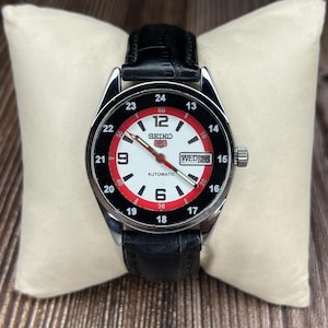 May include: A Seiko automatic wristwatch with a black leather band. The watch face is white with red accents, black numerals, and a day/date display. The watch is displayed on a cream-colored pillow.