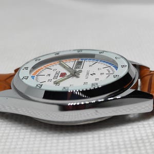 May include: A silver-toned Seiko automatic watch with a white face, featuring black numerals and a day-date display. The watch has a brown leather strap. The watch face has orange and blue accents.