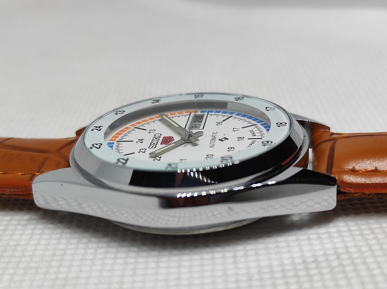 May include: A silver-toned Seiko automatic watch with a white face, featuring black numerals and a day-date display. The watch has a brown leather strap. The watch face has orange and blue accents.