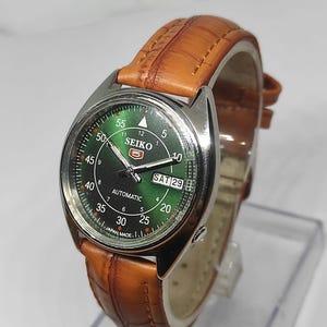 Könnte beinhalten: Eine Seiko 5 Automatik-Uhr mit grünem Zifferblatt und braunem Lederarmband. Das Zifferblatt zeigt den Tag und das Datum mit dem Text "SAT 29". Die Uhr hat ein Edelstahlgehäuse und ist mit "JAPAN MADE" gekennzeichnet.