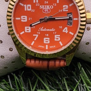 May include: A gold-coloured Seiko 5 automatic watch with an orange dial and a brown leather strap. The watch face displays white numerals and hands, with the text "SEIKO 5" and "Automatic".