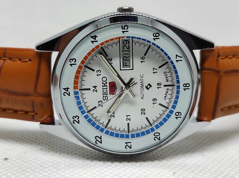 May include: A silver-toned Seiko 5 automatic watch with a white face, featuring a day and date display. The watch has a brown leather band. The face has orange and blue accents, with the day displayed as "FRI 29".
