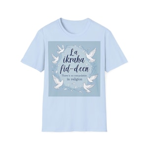 Puede incluir: Camiseta azul claro con un diseño central. El diseño presenta el texto "La ikraha fid-deen" y "There is no compulsion in religion". Palomas blancas y elementos florales rodean el texto, creando una estética pacífica.