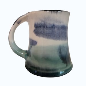 May include: A ceramic mug with a curved handle, featuring a gradient of colors. The mug transitions from a white top to blue and green hues at the base. The design has a watercolor effect, with darker blue patches.