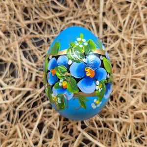 Op de afbeelding: Een handbeschilderd, blauw paasei met een bloemmotief. Het ei heeft blauwe bloemen met groene bladeren en witte accenten, geaccentueerd door een gouden band. Het ei ligt op een stro achtergrond.