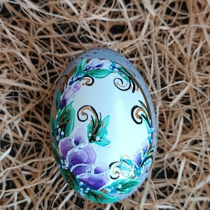 Könnte beinhalten: Ein handbemaltes dekoratives Ei mit floralem Design. Das Ei hat eine weiße Basis mit lila, blauen und grünen Blumen und Blättern. Das Ei ist von einem grauen Rand umgeben und liegt auf einem Strohbett.