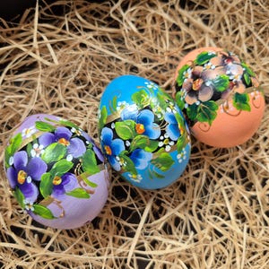 Op de afbeelding: Drie handgeschilderde paaseieren met bloemmotieven liggen op een bedje van stro. De eieren zijn beschilderd in tinten lavendel, blauw en perzik, elk versierd met levendige bloemen en groene bladeren. Het kunstwerk toont ingewikkelde details en een feestelijk thema.