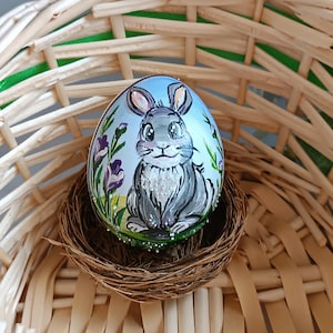 Könnte beinhalten: Handbemaltes Osterei mit einem grauen Hasen vor blauem Himmel und lila Blumen. Das Ei liegt in einem kleinen, braunen Nest in einem geflochtenen Korb. Glitzerakzente verzieren das Ei.