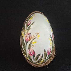 Op de afbeelding: Een handbeschilderd wit ei versierd met roze tulpen en groene bladeren. Het ei is omwikkeld met touw en staat tegen een zwarte achtergrond. Het bloemmotief heeft gele accenten.