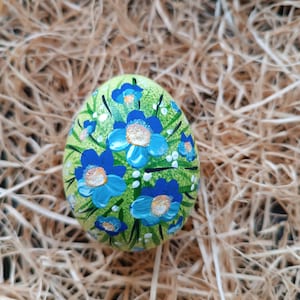 Puede incluir: Un huevo de Pascua pintado a mano con una base verde claro, adornado con flores azules y detalles blancos. El huevo está colocado en un lecho de paja, creando una exhibición rústica y festiva. El huevo es un objeto decorativo.